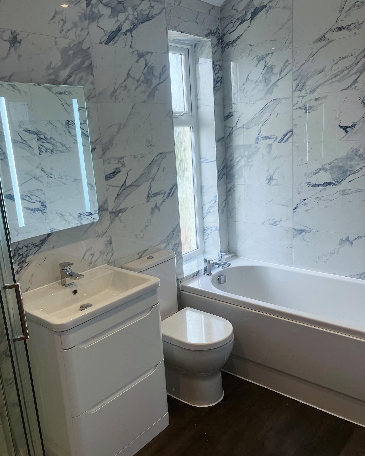 Fitted bathroom with marble-effect wall tiling, a bath under the window and a matching vanity basin
