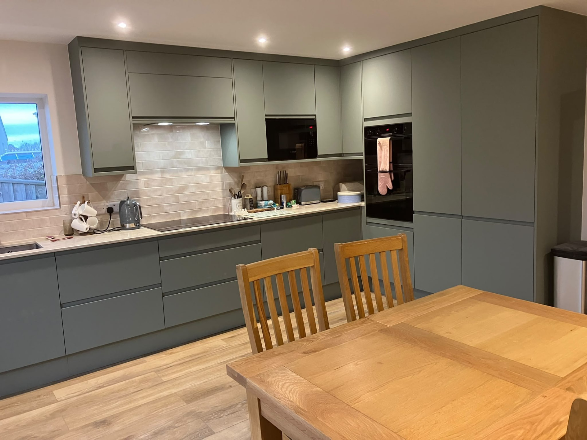 Fitted kitchen with sage green cabinets, tiled splashback and a timber dining table