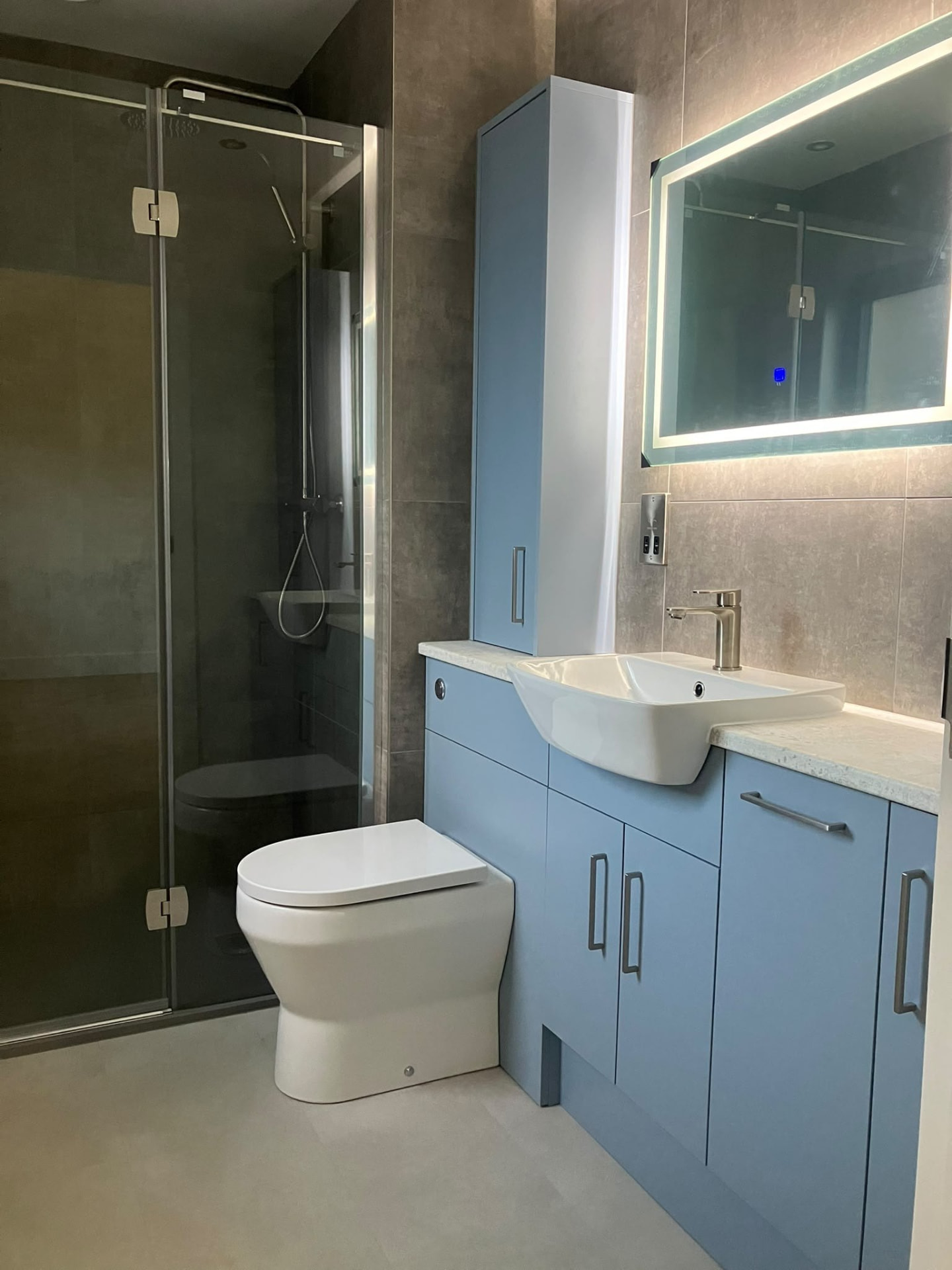 Modern bathroom fit-out with large-format grey tiles, a blue vanity unit and a backlit mirror