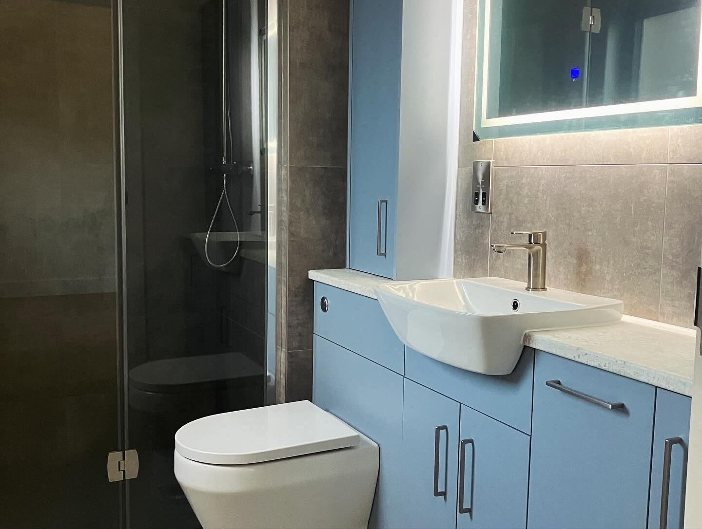 Fitted blue vanity unit and basin in a grey-tiled bathroom with a backlit mirror